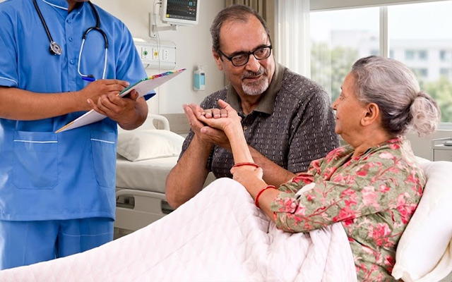 Elderly woman in hospital bed holding hands with her spouse while nurse records vital signs, showing patient-centered care, safe medication management, and emergency readiness in an accredited hospital for senior citizens.